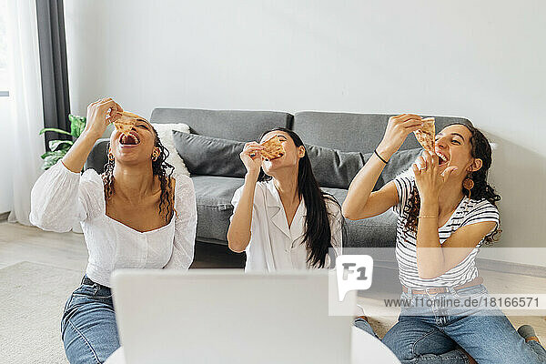 Happy friends eating pizza slices sitting in living room