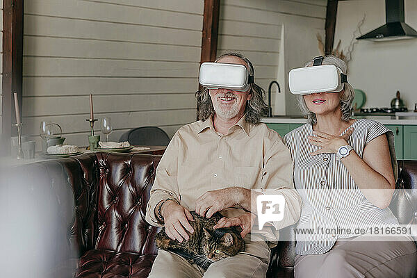 Couple wearing virtual reality simulator glasses sitting with cat on lap at home