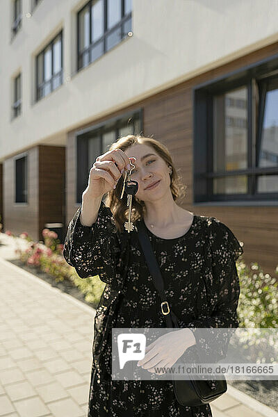 Smiling young woman showing house keys standing in front of building