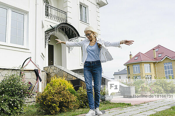 Smiling woman with arms outstretched skateboarding in front of house