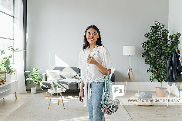 Smiling woman carrying mesh bag with vegetables in living room