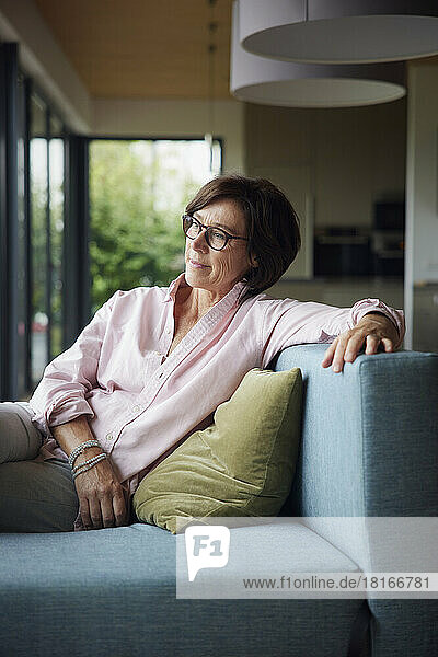 Thoughtful senior woman with eyeglasses sitting on sofa at home