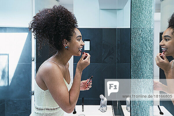 Glückliche Frau mit lockigem Haar  die zu Hause im Badezimmer Lippenstift aufträgt