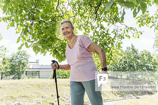 Lächelnde ältere Frau geht mit Wanderstock am Baum entlang