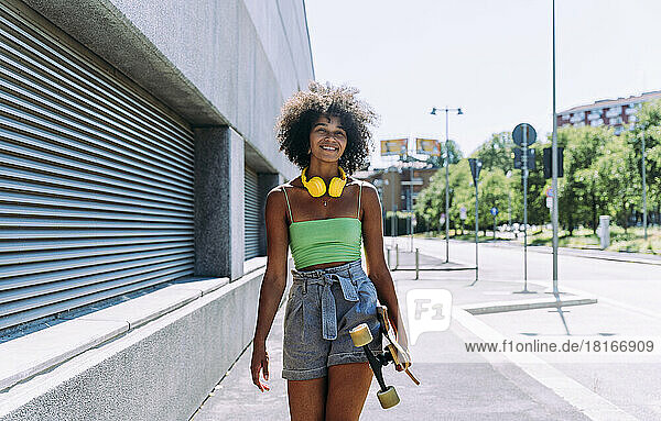 Smiling young woman with skateboard walking on footpath