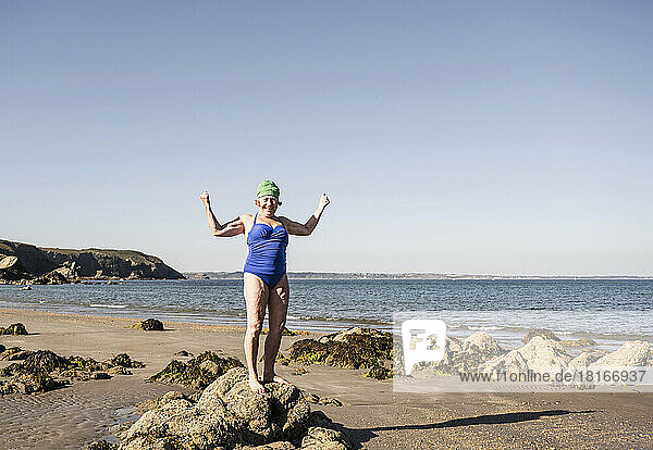 Ältere Frau steht auf einem Felsen und lässt am Strand ihre Muskeln spielen