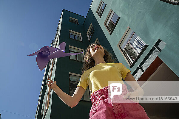 Smiling woman with hand in pocket holding pinwheel toy standing near building