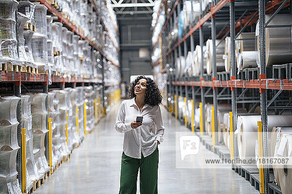 Smiling businesswoman holding smart phone standing with hand in pocket at factory