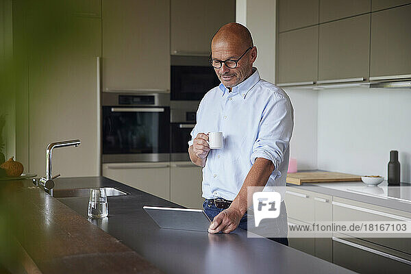 Smiling man with coffee cup using tablet PC standing at kitchen island
