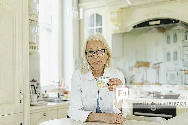 Senior woman holding cup leaning on kitchen island at home