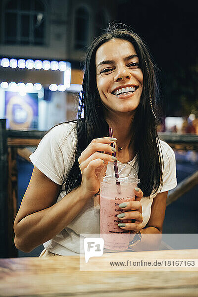 Happy woman holding smoothie sitting at table in sidewalk cafe