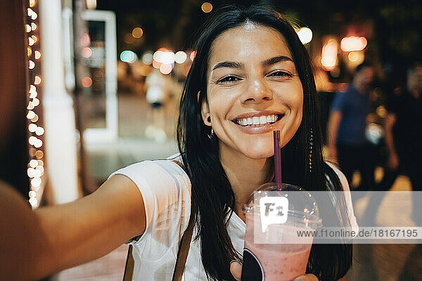 Happy beautiful woman with smoothie taking selfie at night