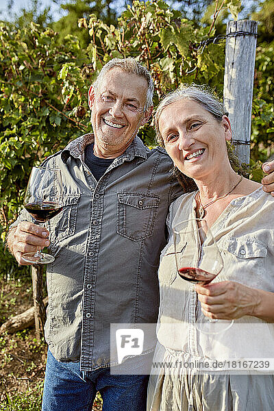 Smiling mature couple holding wineglasses in front of vineyard on sunny day