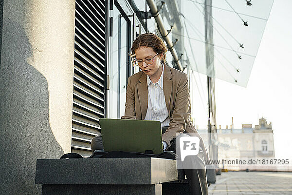 Young businesswoman using laptop on bench by office building