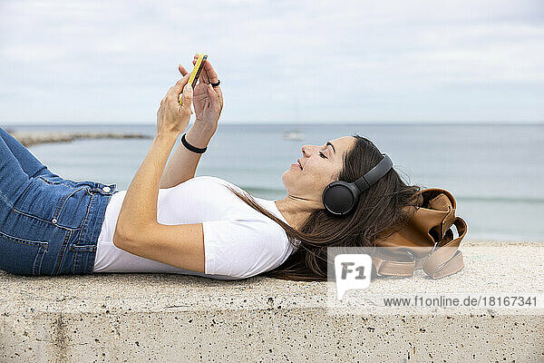Lächelnde Frau  die an der Wand liegt und über kabellose Kopfhörer Musik hört und ihr Smartphone benutzt