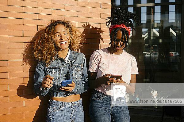 Young woman laughing by friend using mobile phone on sunny day