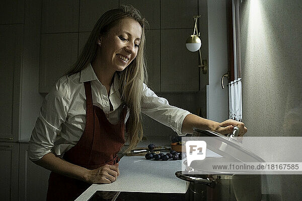 Lächelnde Frau öffnet den Deckel eines Topfes in der Küche