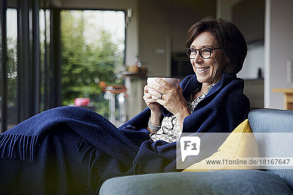 Happy senior woman wrapped in blanket sitting with coffee cup on sofa at home