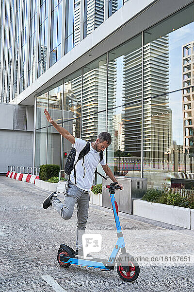 Man with hand raised balancing on electric push scooter in front of buildings