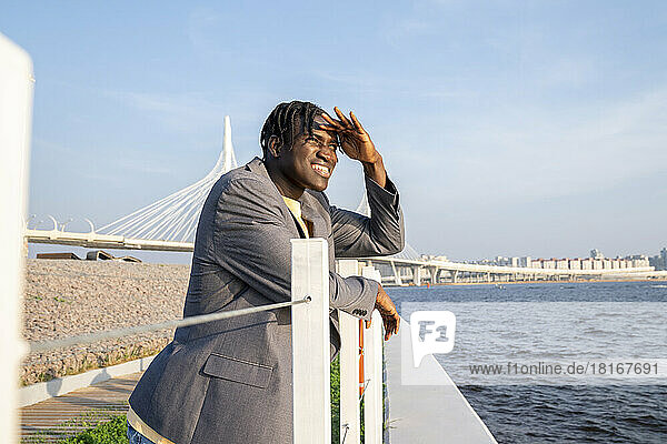Businessman shielding eyes and leaning on railing by sea