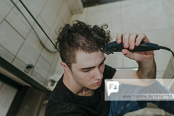 Sad young man with electric razor shaving his own hair in bathroom