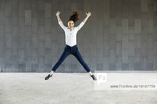 Carefree woman with arms raised jumping in front of wall