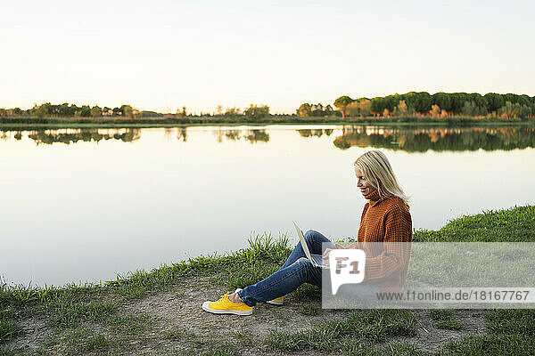 Smiling freelancer working on laptop sitting near lake