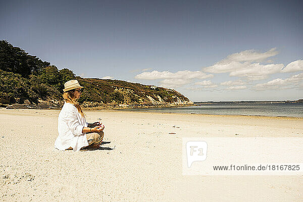 Mature woman wearing hat practicing meditation at beach on sunny day
