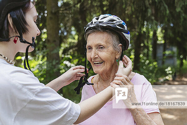 Great granddaughter helping senior woman wearing bicycle helmet in park