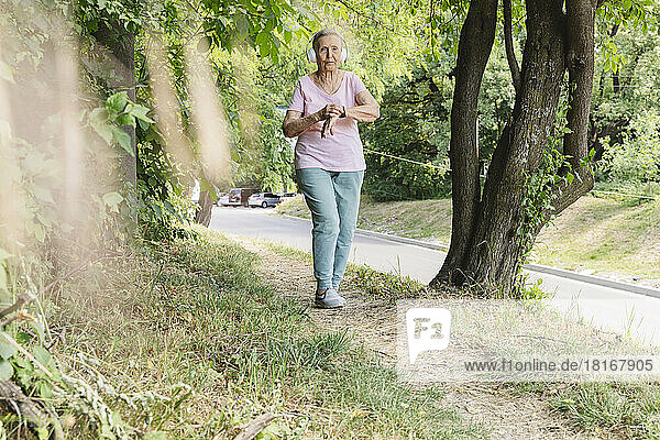 Active senior woman wearing wireless headphones walking by tree in park