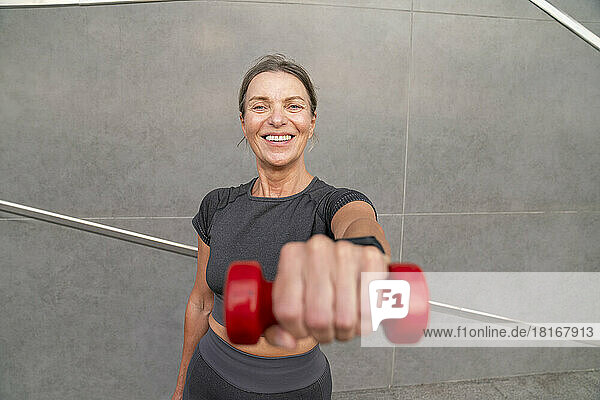 Happy woman showing red dumbbell in front of wall