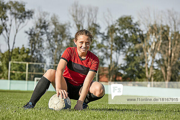 Happy girl with soccer ball crouching on field on sunny day