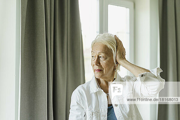 Senior woman touching white hair near curtain at home