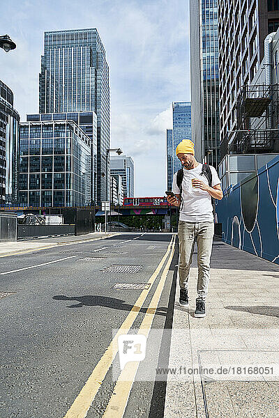Mann benutzt Smartphone und geht an einem sonnigen Tag in der Stadt auf dem Fußweg spazieren