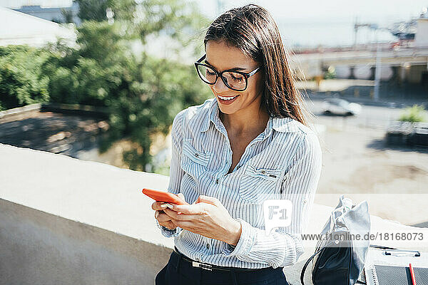 Happy businesswoman using smart phone leaning on wall