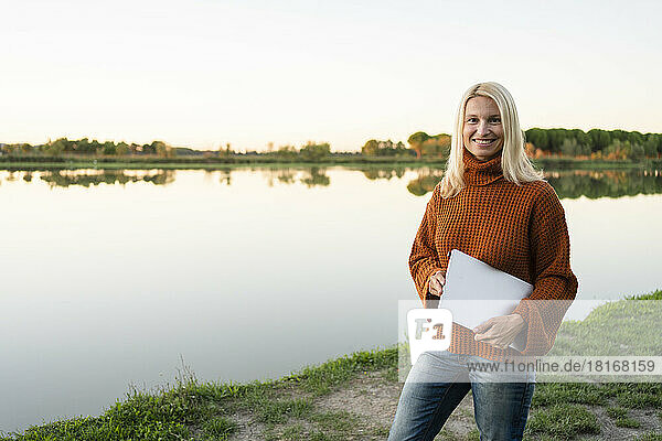 Smiling mature freelancer with laptop standing near lake
