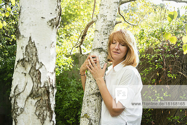 Mature woman with eyes closed embracing tree trunk in garden