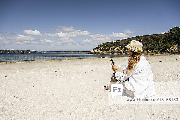 Reife Frau benutzt an einem sonnigen Tag ihr Mobiltelefon am Strand