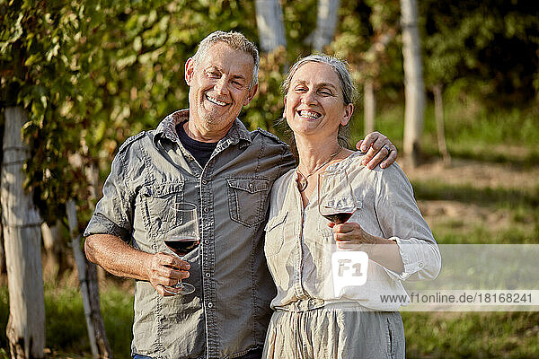 Happy mature couple holding red wineglasses in front of vineyard