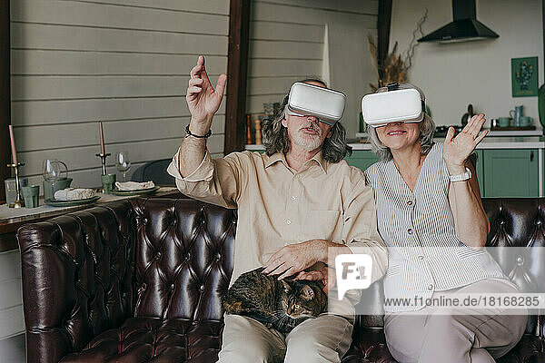 Älteres Paar mit VR-Brille gestikuliert mit Katze auf dem Schoß zu Hause