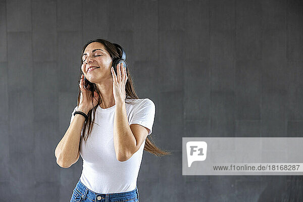 Smiling woman enjoying music through wireless headphones in front of gray wall