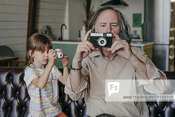 Enkelin mit Spielzeugkamera und Großvater mit Vintage-Kamera fotografieren zu Hause