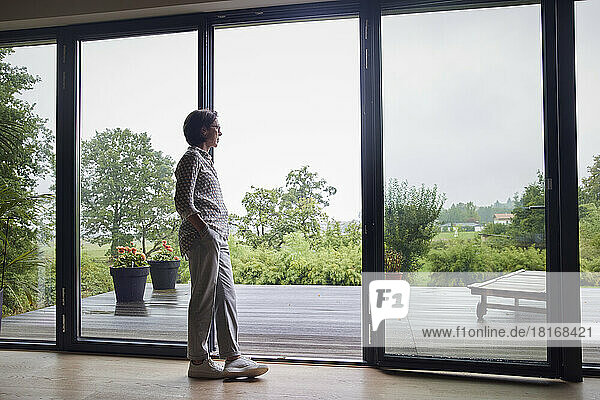 Thoughtful senior woman leaning on glass door at home