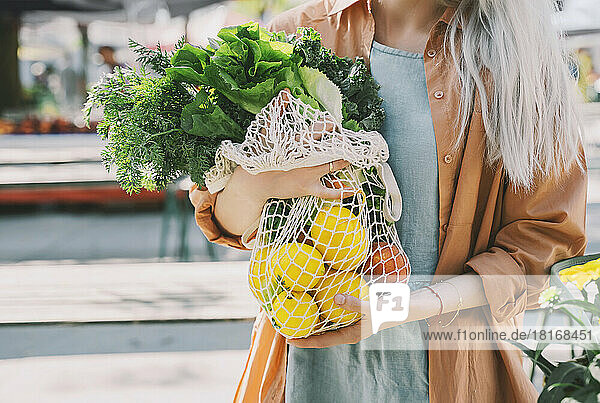 Customer holding leafy vegetables and lemons in mesh bag at local market