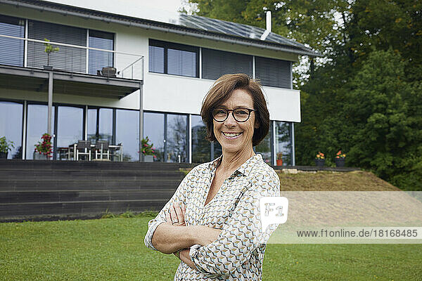 Happy senior woman with arms crossed standing in front of house