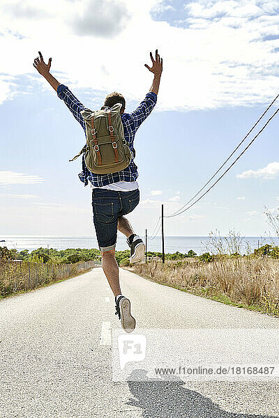 Man wearing backpack jumping with arms raised on sunny day