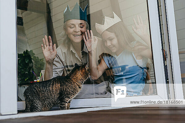 Glückliche Großmutter und Enkelin mit Krone und Blick auf die Katze durch das Glasfenster