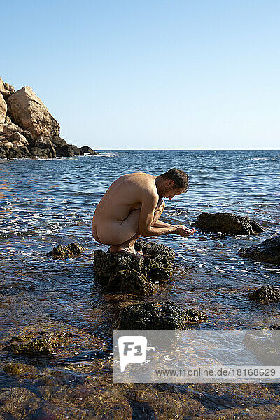 Nackter Mann in hockender Position auf einem Felsen im Meer