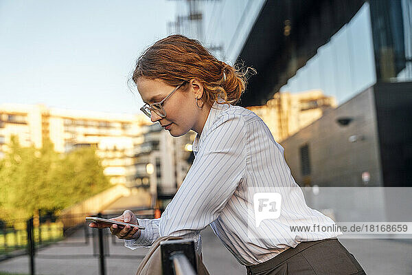 Young redhead businesswoman using mobile phone leaning on railing