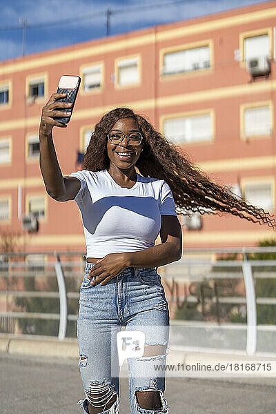 Glückliche junge Frau mit zerzausten Haaren  die auf dem Fußweg ein Selfie mit dem Smartphone macht
